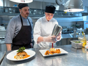 A chef's assistant pours a sauce over a dish of roasted carrots; the chef supervises.