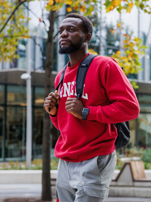 Student in a red Cornell sweatshirt walking outdoors with fall foliage in the background