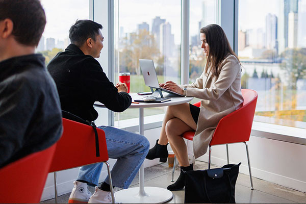 Two Cornell students collaborating at a small table near floor-to-ceiling windows with a city skyline behind them