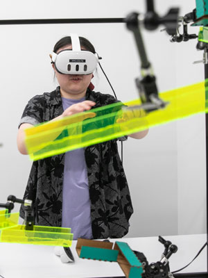 Cornell student wearing a VR headset interacting with a virtual object in a lab surrounded by robotic equipment.
